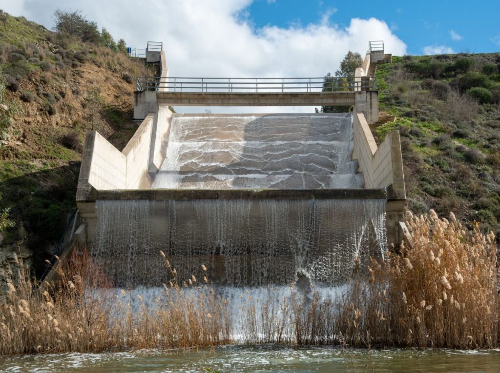 Water flowing over a concrete dam spillway between grassy hills under a blue sky.