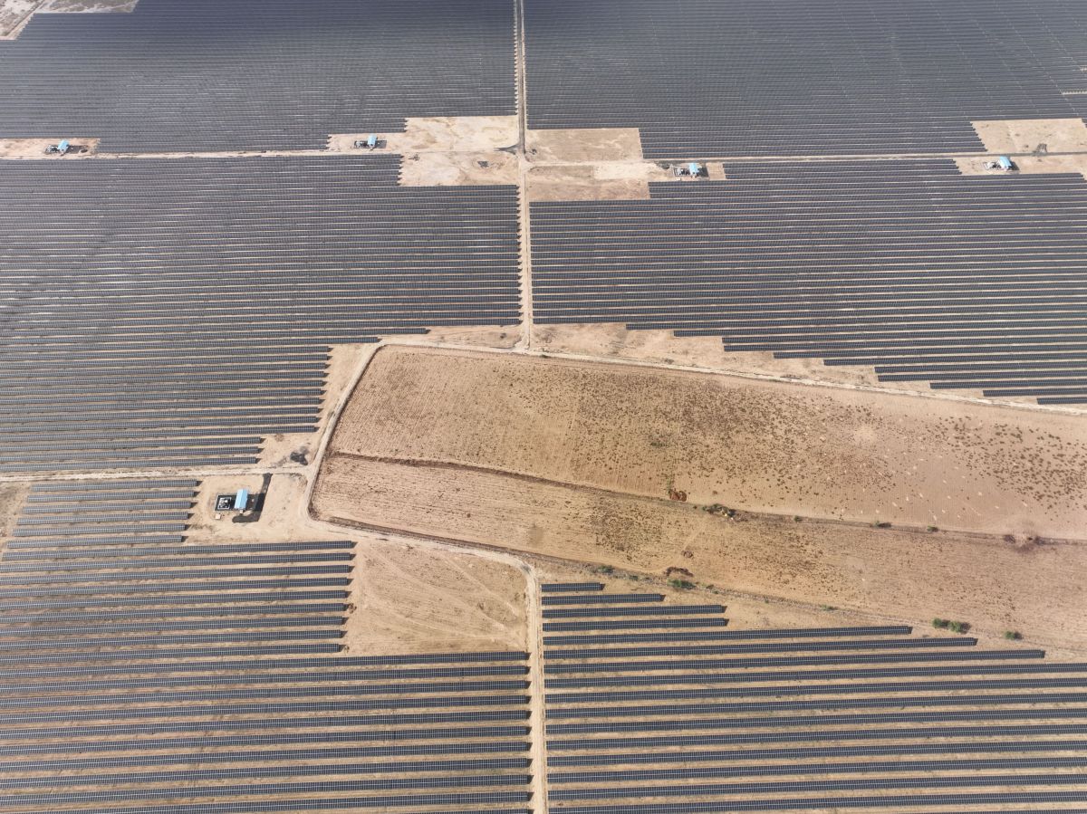 Aerial view of a large solar farm with long rows of dark solar panels arranged in a grid across a dry sandy landscape. Dirt access roads divide the panels into sections with small utility structures visible at intersections.