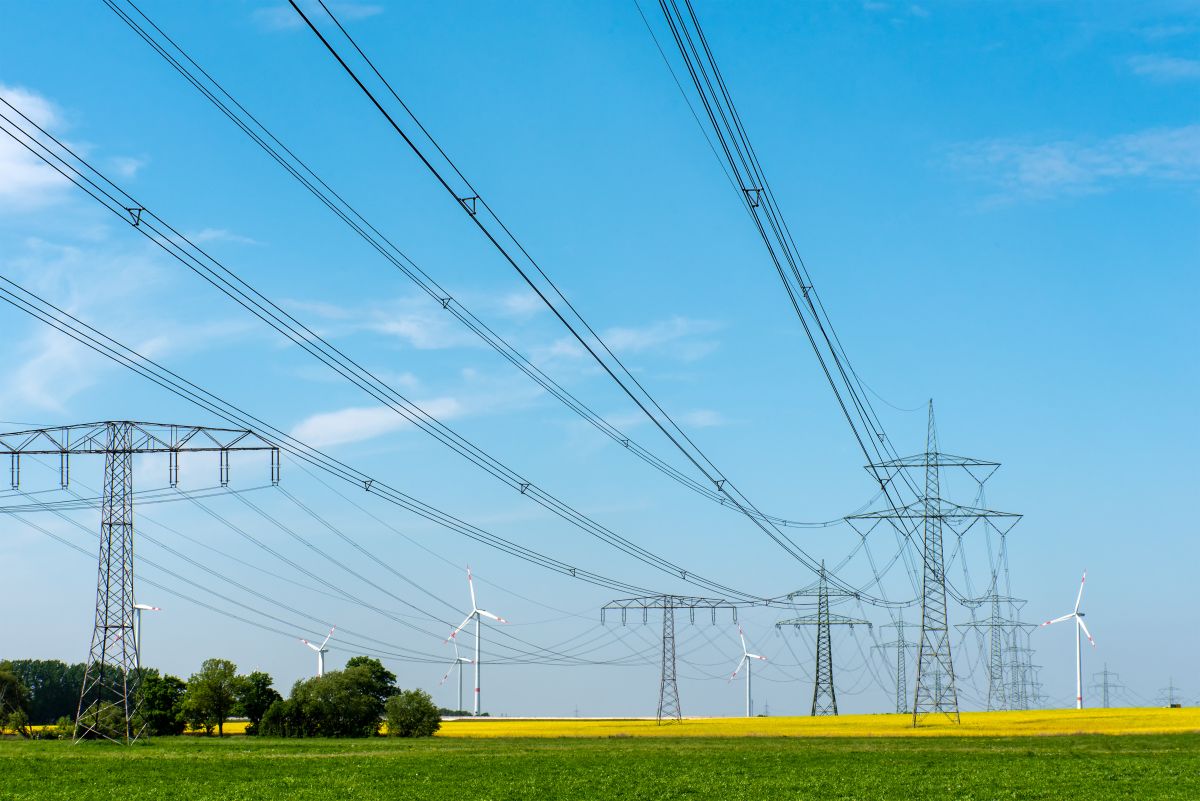High voltage transmission lines and towers stretching across a green and yellow field with wind turbines in the background under a clear blue sky, representing electricity transmission infrastructure and cross border energy flow.
