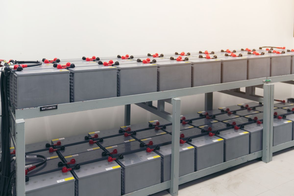 Rows of large industrial battery packs arranged on metal shelving in a power plant battery room, connected by cables and terminals for electricity storage and backup supply.