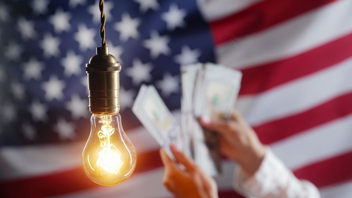 A glowing Edison light bulb hanging in front of a blurred American flag while a person counts dollar bills in the background, illustrating rising electricity costs and energy expenses.