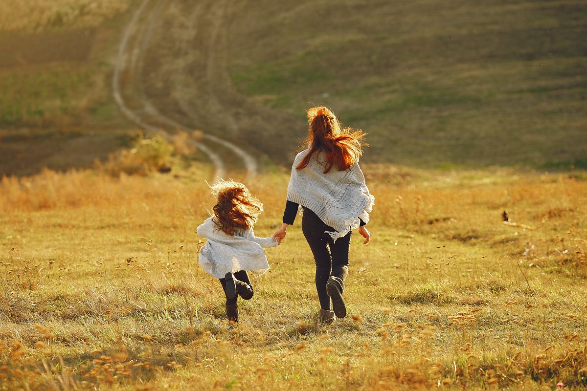 Mother with little daughter playing in a autumn field A woman and a young girl run hand in hand through a golden autumn field with dry grass and a winding path stretching into the distance.