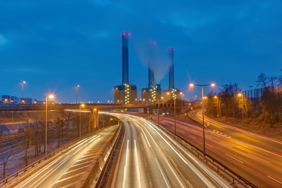 FERC Rejects MISO Plan to Speed Generation Interconnection - Certrec A power plant with tall smokestacks emitting smoke in the distance while highway traffic creates light trails in the foreground at night, showing electricity generation supporting urban infrastructure.