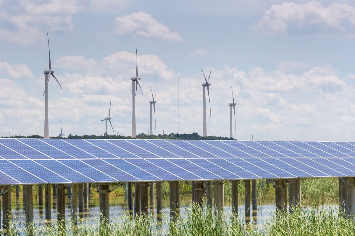 Solar panels mounted above a grassy waterlogged field with wind turbines spinning in the background, showing a combined wind and solar renewable energy site.