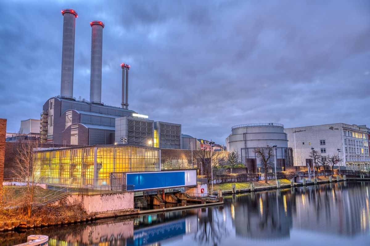 A power plant with multiple smokestacks beside a calm body of water at dusk, with lights glowing from the building and reflecting on the surface, showing electricity generation infrastructure near an urban area.