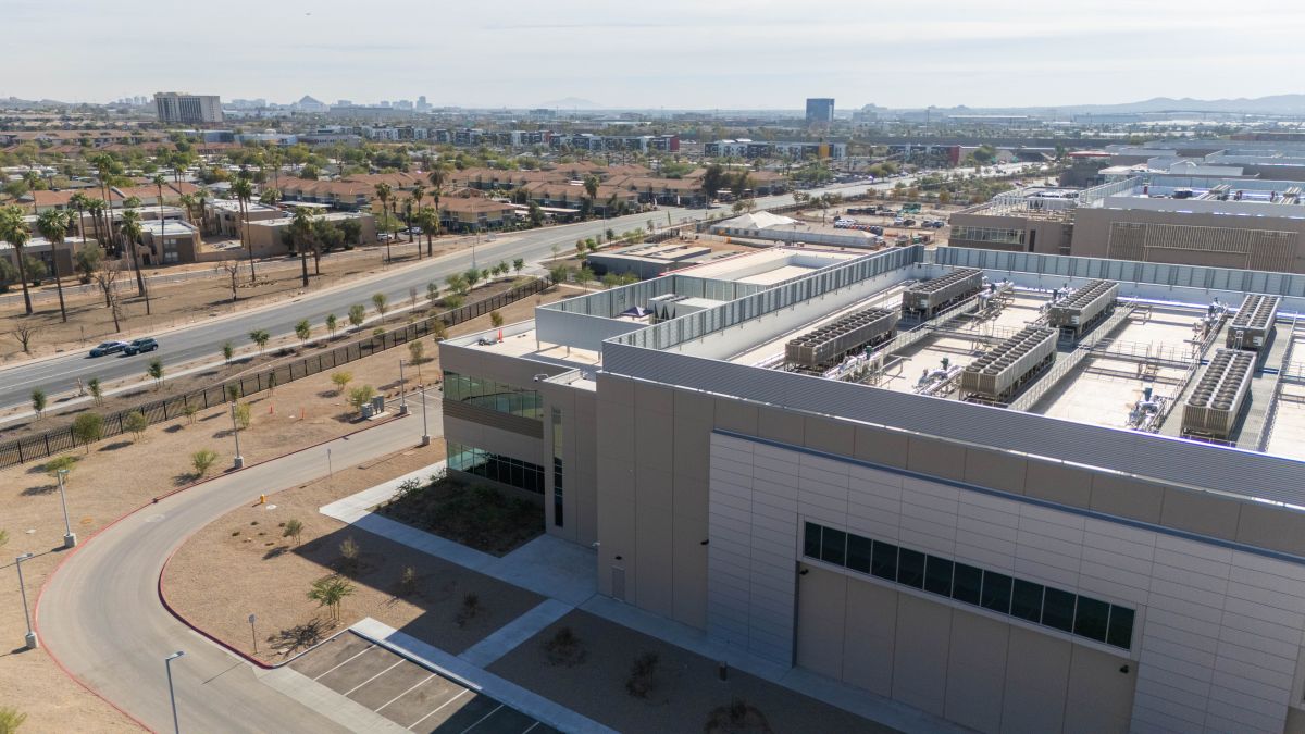 Aerial view of a modern industrial facility with rooftop equipment and cooling units, located near roads and surrounded by a suburban urban area with houses and low rise buildings.
