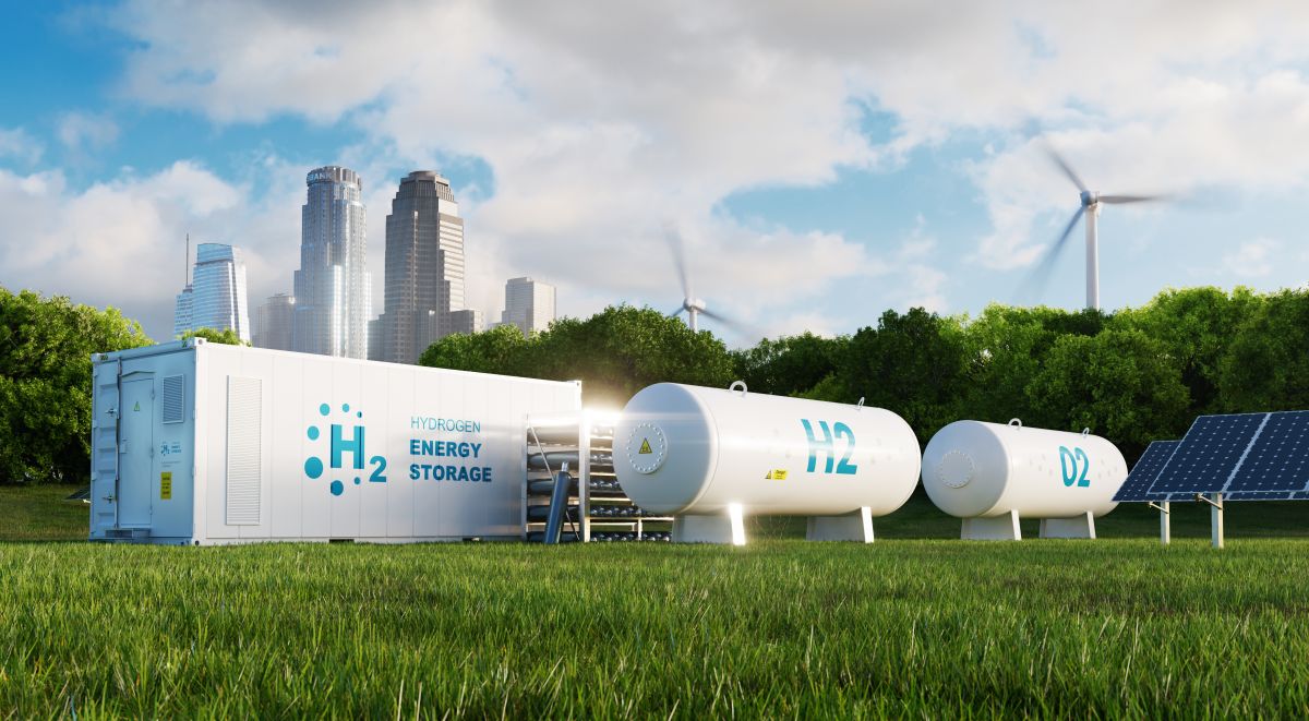 Hydrogen energy storage facility set on a grassy field with a city skyline in the background. The image shows a white H₂ storage unit alongside cylindrical tanks labeled H2 and O2, surrounded by solar panels and wind turbines, illustrating a clean energy ecosystem.