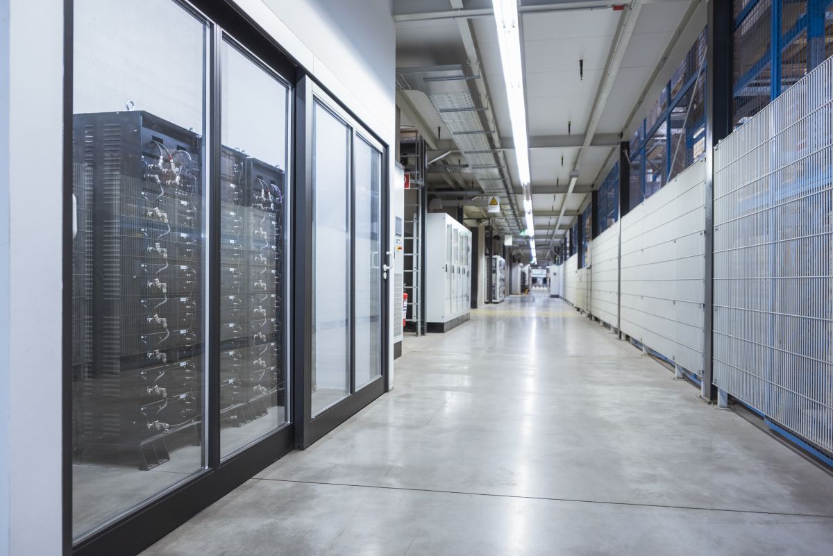 A factory corridor with rows of industrial battery storage units enclosed behind glass panels, showing buffer energy storage systems used to manage power supply and demand.