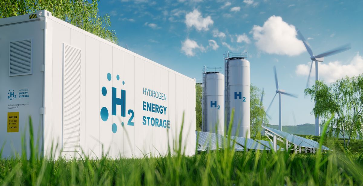 A clean energy facility featuring a white hydrogen energy storage container labeled "H₂ Hydrogen Energy Storage" sits in a grassy field. In the background, wind turbines, solar panels, and tall H₂ storage tanks highlight a renewable energy system under a bright, partly cloudy sky.