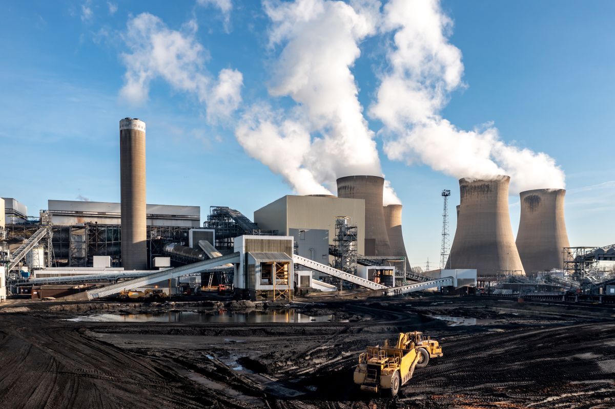 Heavy machinery loading coal into a coal-fired power plant, with large cooling towers and smoke rising under a clear blue sky.