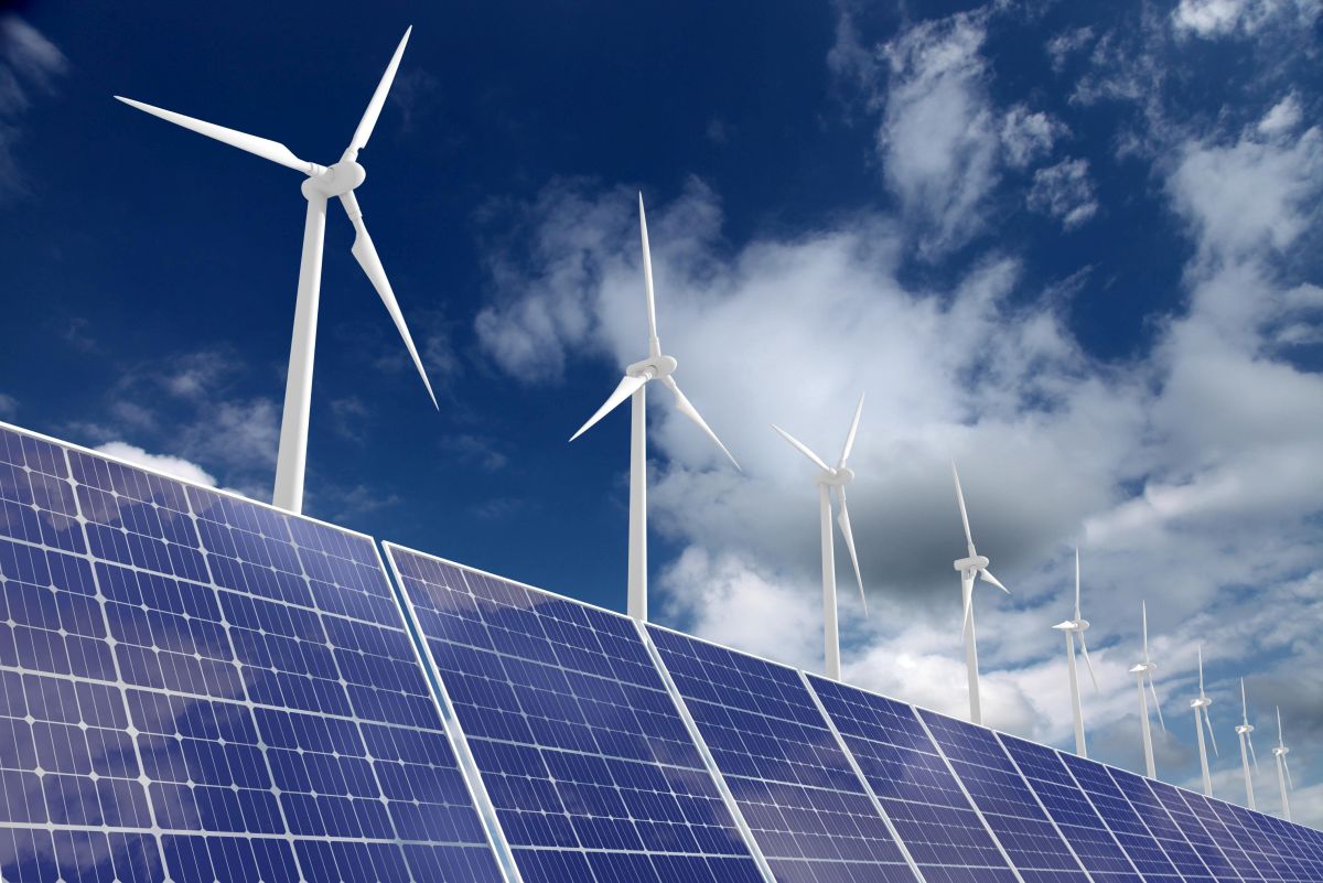 Solar panels in the foreground with wind turbines under a bright blue sky with scattered clouds.