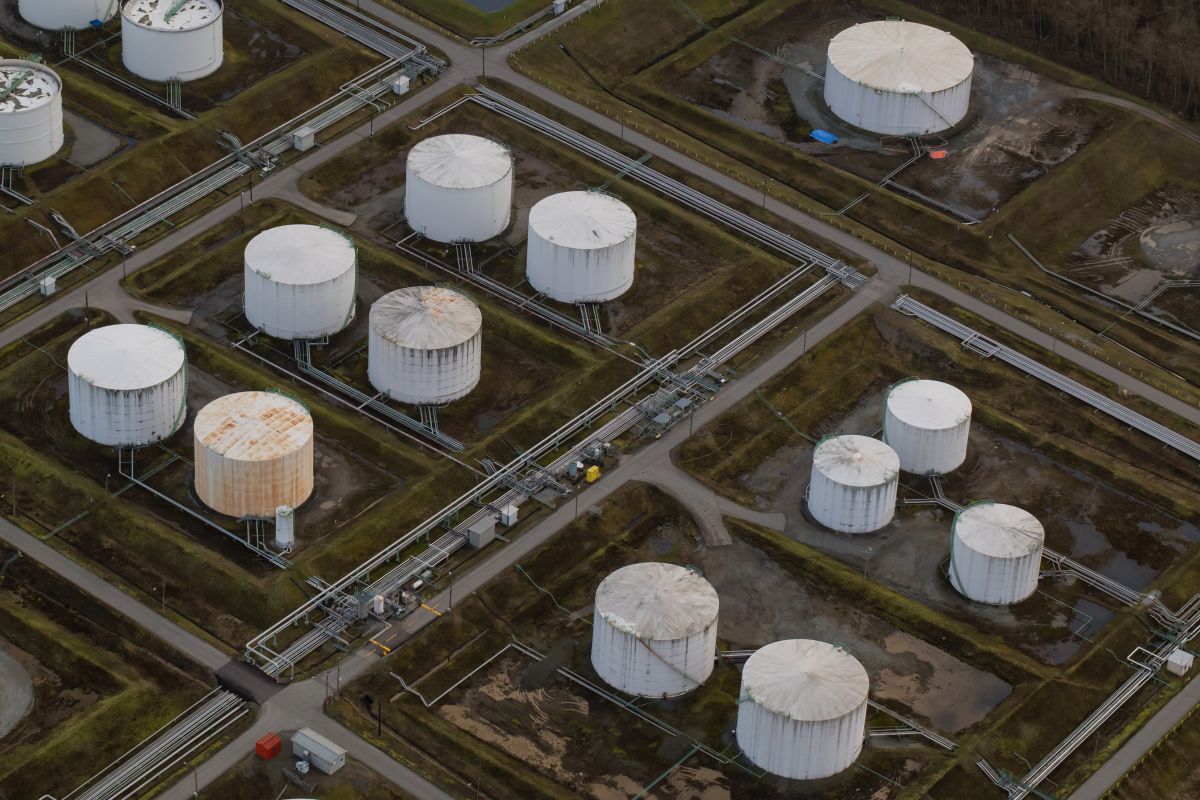 Aerial view of an oil refinery facility with large storage tanks connected by pipes, set against a green industrial landscape.