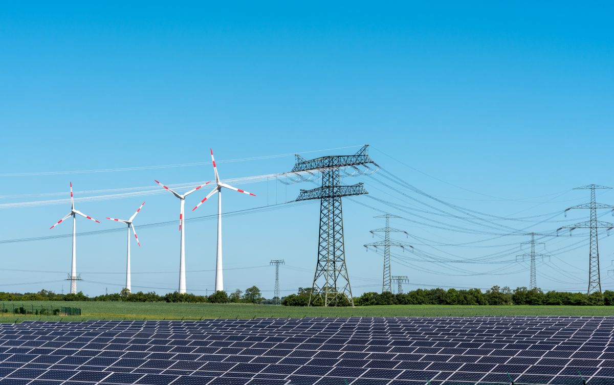 Solar panels and wind turbines beneath transmission lines in a clear blue sky.