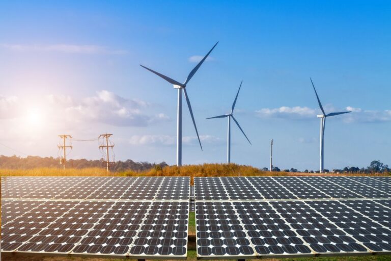 Solar panels in the foreground with wind turbines in the background, under a clear blue sky with sunlight.