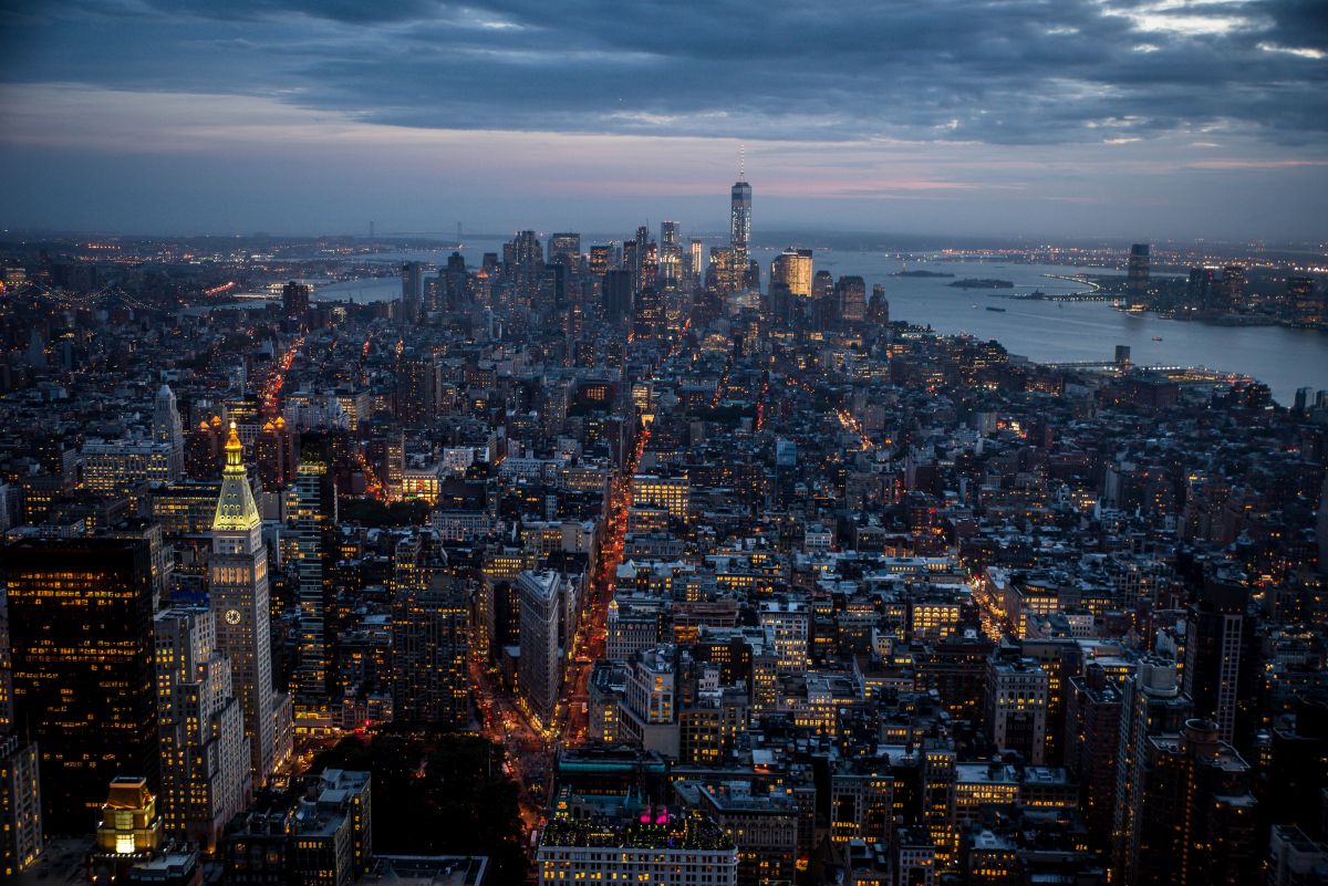 Manhattan skyline at dusk with city lights glowing beneath dark clouds and the river in the background.
