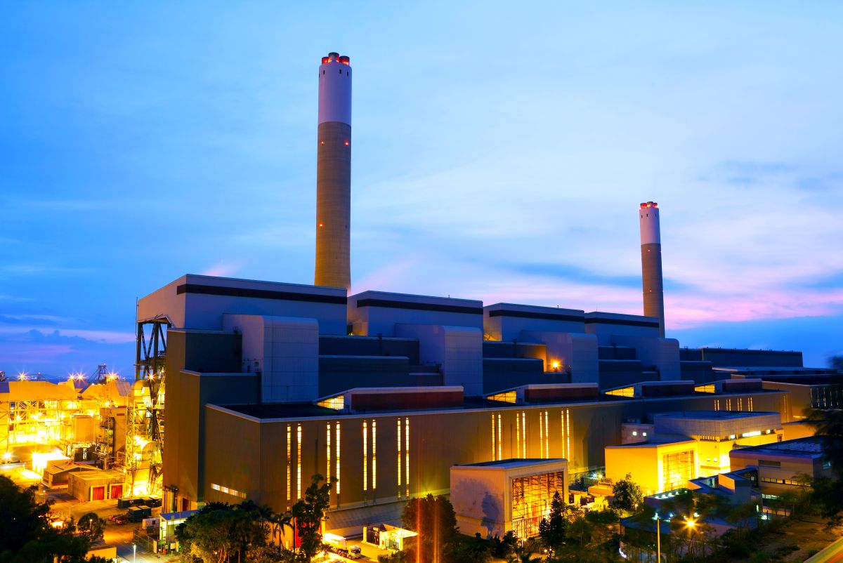 Power plant at dusk with lit buildings and two tall smokestacks against a blue evening sky.