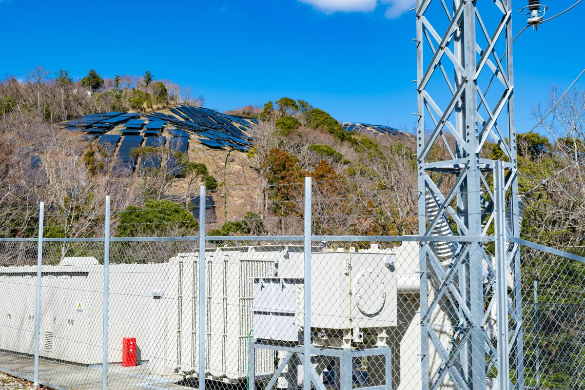 Battery storage units secured behind a fence at a grid facility with solar panels on a hillside in the background. The image highlights large scale battery storage supporting grid reliability and renewable energy integration.