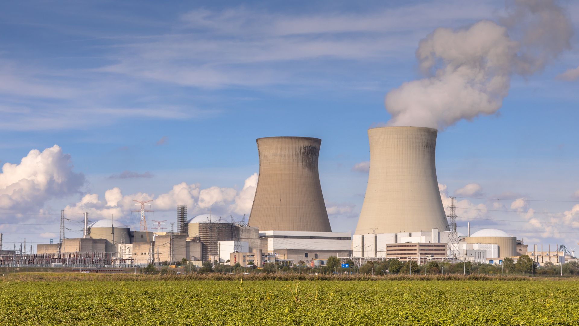 This image shows a nuclear power plant with two large cooling towers emitting steam into the sky. The plant is situated in a rural area with fields in the foreground, and various power lines and infrastructure are visible in the scene. The cloudy sky further enhances the industrial atmosphere of the power plant.
