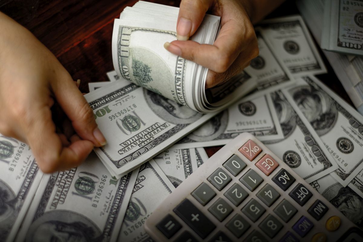 A close-up of a person counting a stack of $100 US dollar bills, with a calculator placed next to the money.