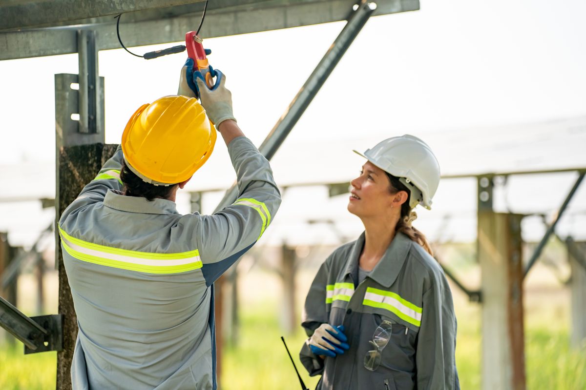 Engineer in a yellow hard hat using an energy measurement tool to test wiring beneath photovoltaic panels while a colleague observes. The image highlights inspection and performance testing of photovoltaic modules for energy production.