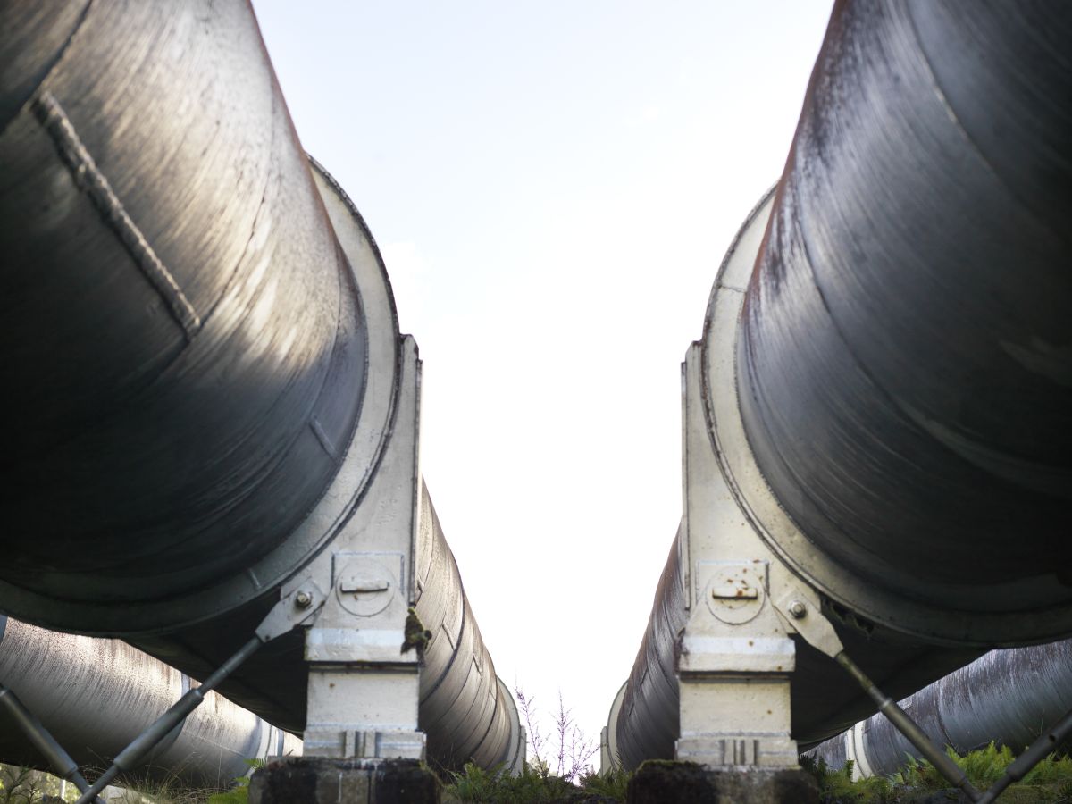 Low-angle view between two large hydroelectric industrial pipes, with supports visible in the foreground and sky in the background.