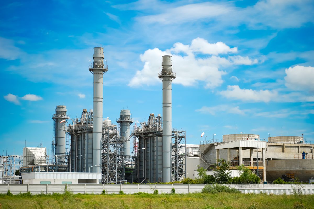 A gas power plant with large smokestacks and industrial structures under a blue sky, featuring green grass in the foreground.