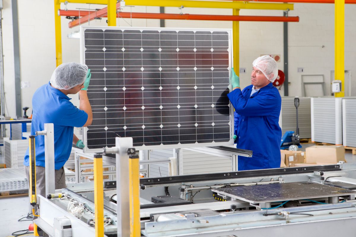 Two workers in a solar panel manufacturing facility carefully handle a large solar panel, with one worker adjusting its position and the other supporting it. Both are wearing protective gear, including gloves and hairnets, in a clean, well-lit industrial setting.