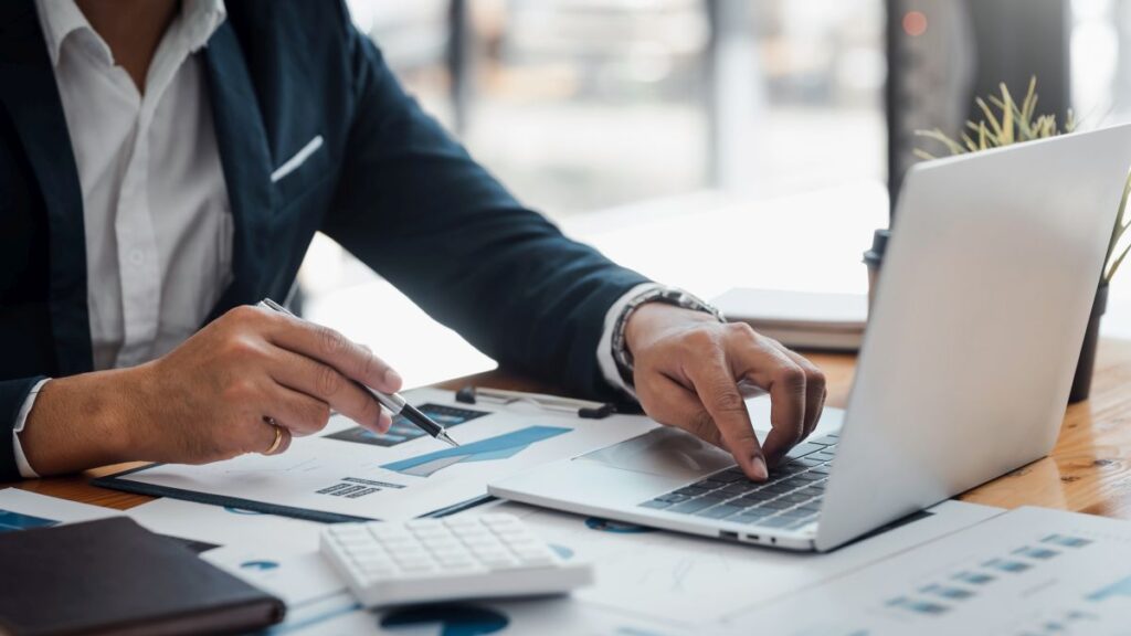 The image shows a businessman using a laptop while sitting at a table in an office, with documents and a pen in front of him.