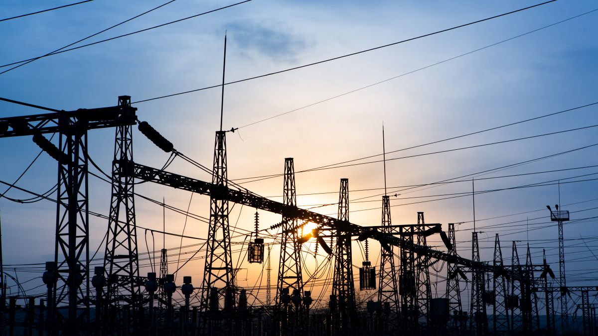 The image shows a silhouette of a high-voltage transformer station at sunset. The power lines and electrical structures are outlined against the colorful sky, showcasing the complexity of the electrical transmission network.