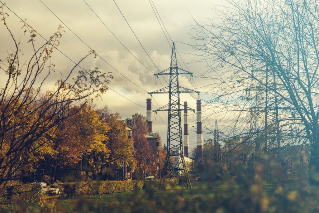 Power lines and electricity pylons with a backdrop of industrial chimneys and autumn trees, under a cloudy sky.
