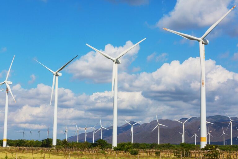Wind turbines standing across an open landscape with mountains in the background under a bright blue sky. The image highlights utility scale wind energy generation in a rural setting.