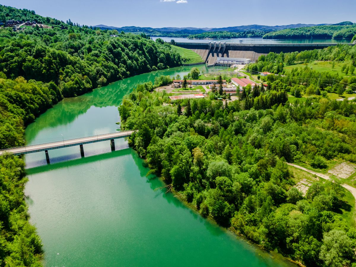 The image shows a hydropower plant on Solina Lake in Poland, surrounded by green forests and a bridge over the lake.