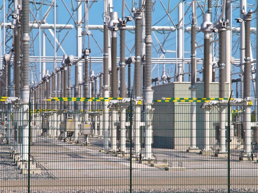 A detailed view of an electrical substation, showing transmission lines, transformers, and metal fences, with clear blue skies in the background.