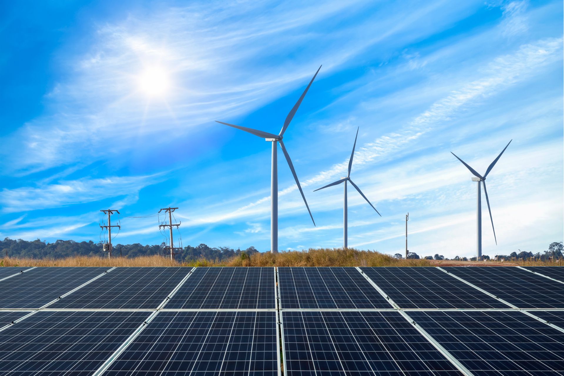 Solar panels in the foreground with wind turbines turning under a bright blue sky. The image highlights photovoltaic modules and wind energy working together at a renewable power plant.