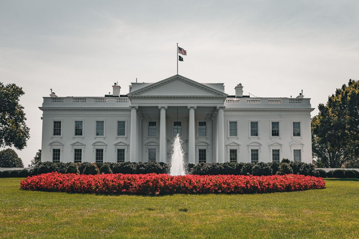 The White House, with a fountain in the foreground and a garden of red flowers, against a bright sky.