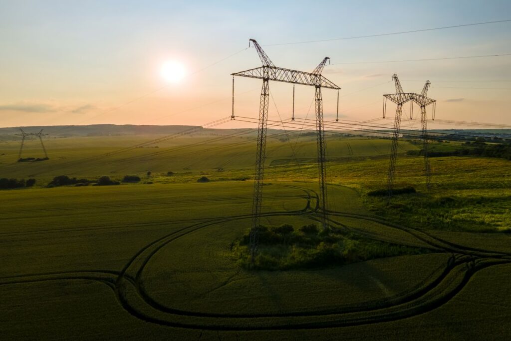 Silhouette of high-voltage power towers and electric power lines against a sunrise, with a green field in the foreground.