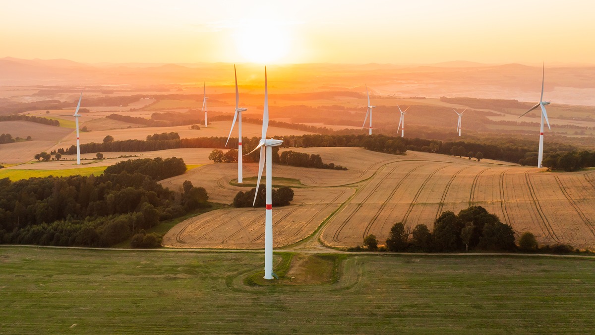 A wide aerial view of a rural landscape at sunset with multiple white wind turbines scattered across golden farmland and green fields. The soft orange glow of the sun highlights the clean energy infrastructure and rolling hills in the distance, symbolizing sustainable energy efforts.
