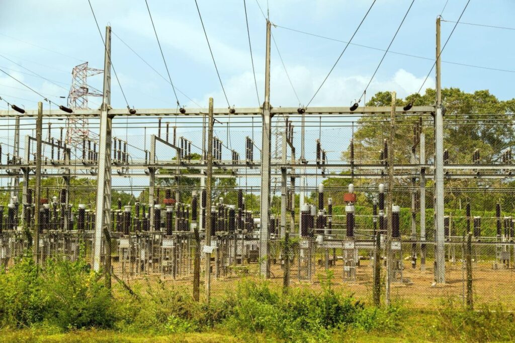 Outdoor electrical substation surrounded by fencing, with transformers, insulators, and overhead power lines set against a backdrop of trees and a blue sky. The image supports content related to grid infrastructure and NERC compliance requirements.