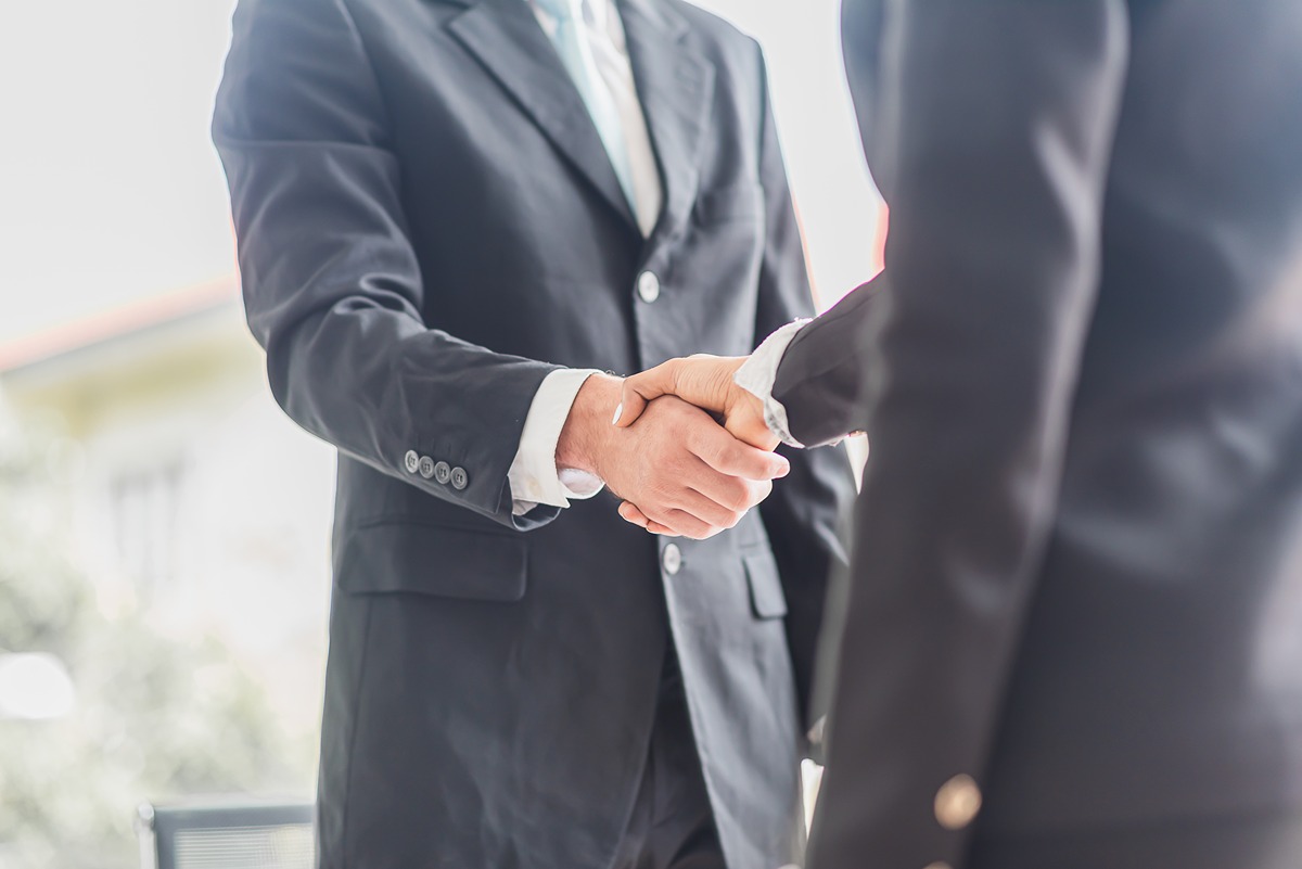 Two people in business suits shaking hands in a bright office setting, symbolizing a formal partnership or agreement. The image supports the announcement of collaboration agreements by the Energy Department with 24 organizations.