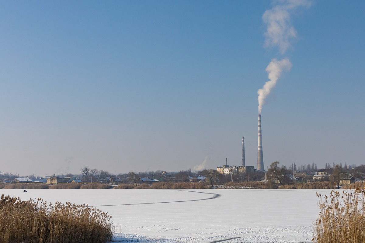 A coal power plant with tall smokestacks emits white smoke into a clear blue sky during winter. The foreground shows a frozen, snow-covered landscape with dry reeds, indicating a cold climate.