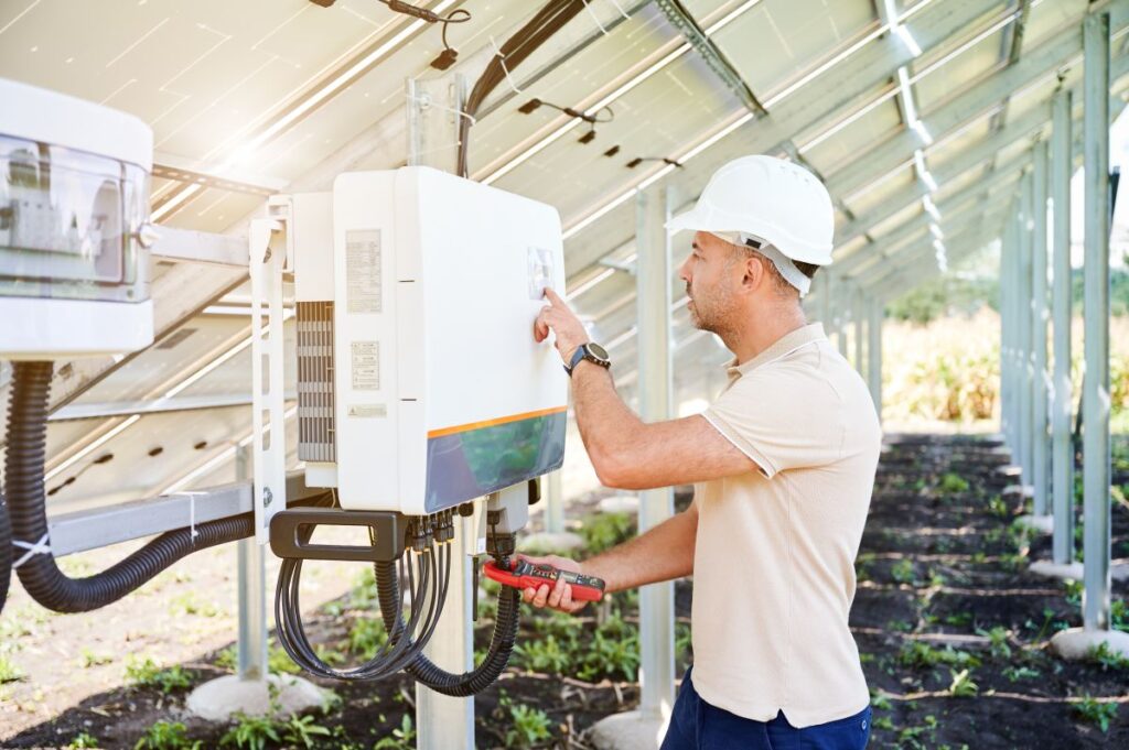 A professional worker wearing a white hard hat and beige shirt checks a solar inverter mounted on a metal support under a row of solar panels. He holds a voltage tester in one hand while pressing a button on the inverter with the other. The setting is an outdoor solar energy installation with visible wiring and structural supports.