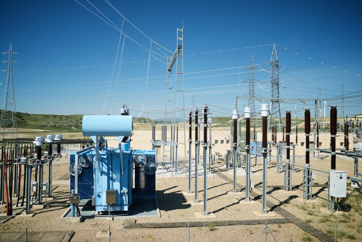 Electrical substation featuring a large blue transformer, circuit breakers, and high-voltage power lines, set in a rural area under a clear sky. The image represents infrastructure relevant to NERC standards for GMD (geomagnetic disturbance) event compliance and planning challenges.
