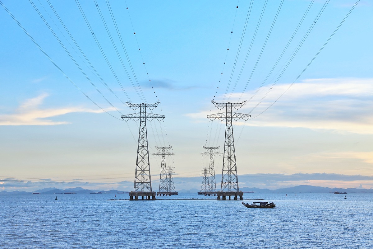 Series of high-voltage power transmission towers stretching across a body of water, under a blue sky with scattered clouds. A small boat is seen in the foreground, and distant landmasses and additional towers are visible in the background, illustrating large-scale electricity infrastructure.