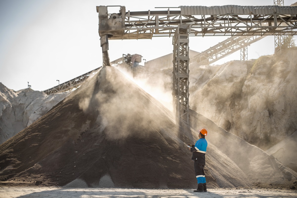Worker in a hard hat and safety gear stands at the base of a large coal pile under a conveyor system at an active coal facility. Dust rises as coal is dumped from overhead machinery, illustrating operations at a Colorado coal unit ordered to remain running.