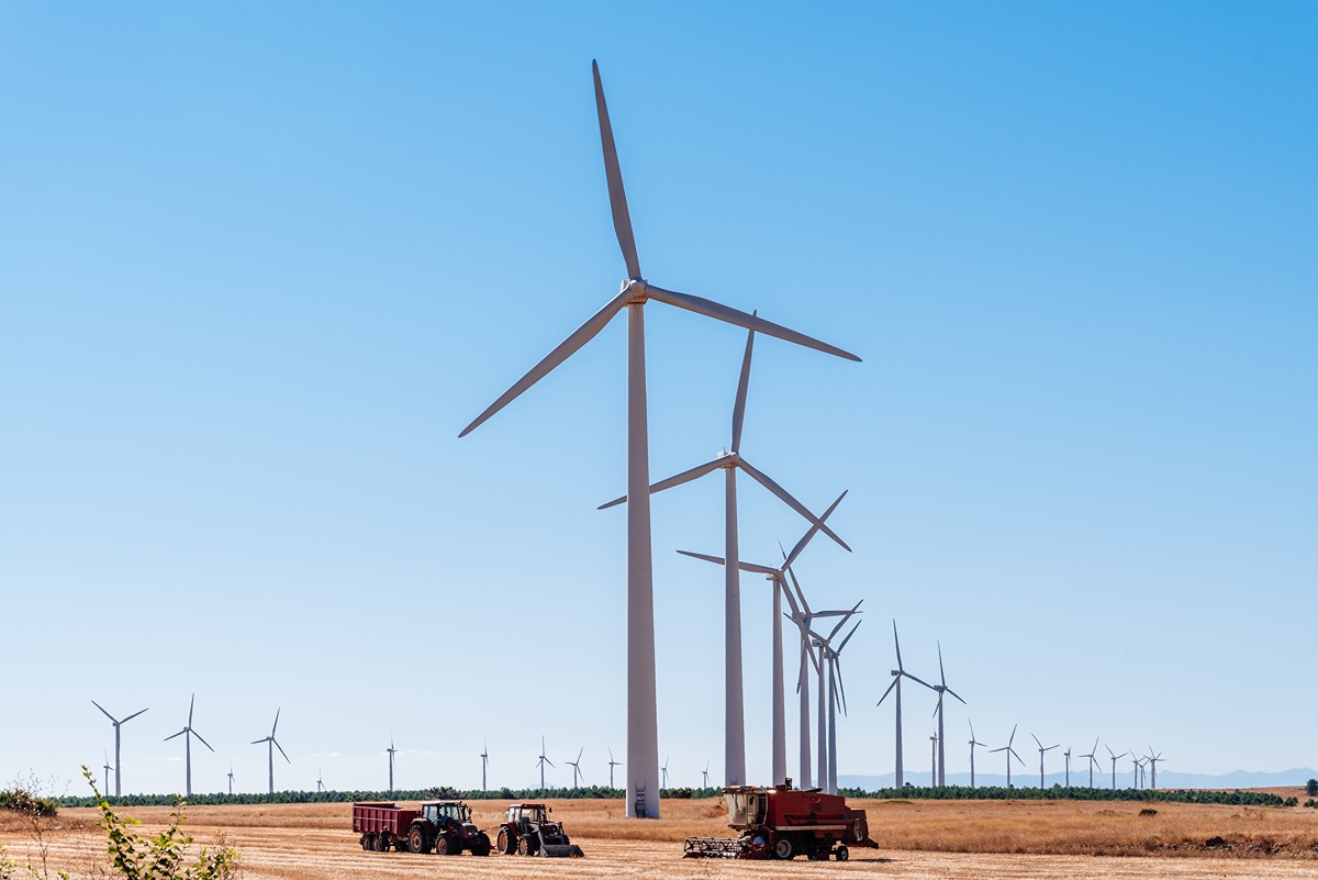Wind farm with rows of large wind turbines stretching across a dry field under a clear blue sky, with construction and farming vehicles in the foreground. The image represents the installation of Dominion’s first wind turbine tower off the Virginia Beach coast.