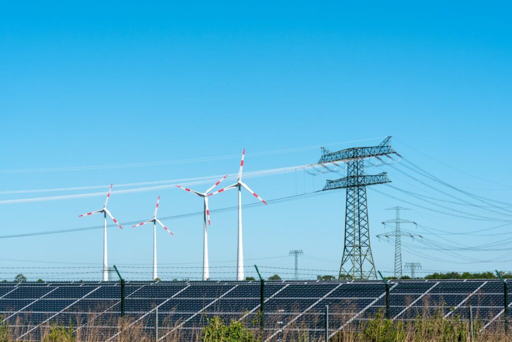 A renewable energy facility featuring solar panels in the foreground, several wind turbines with red tips in the middle ground, and tall electrical transmission towers and power lines in the background, all under a clear blue sky.