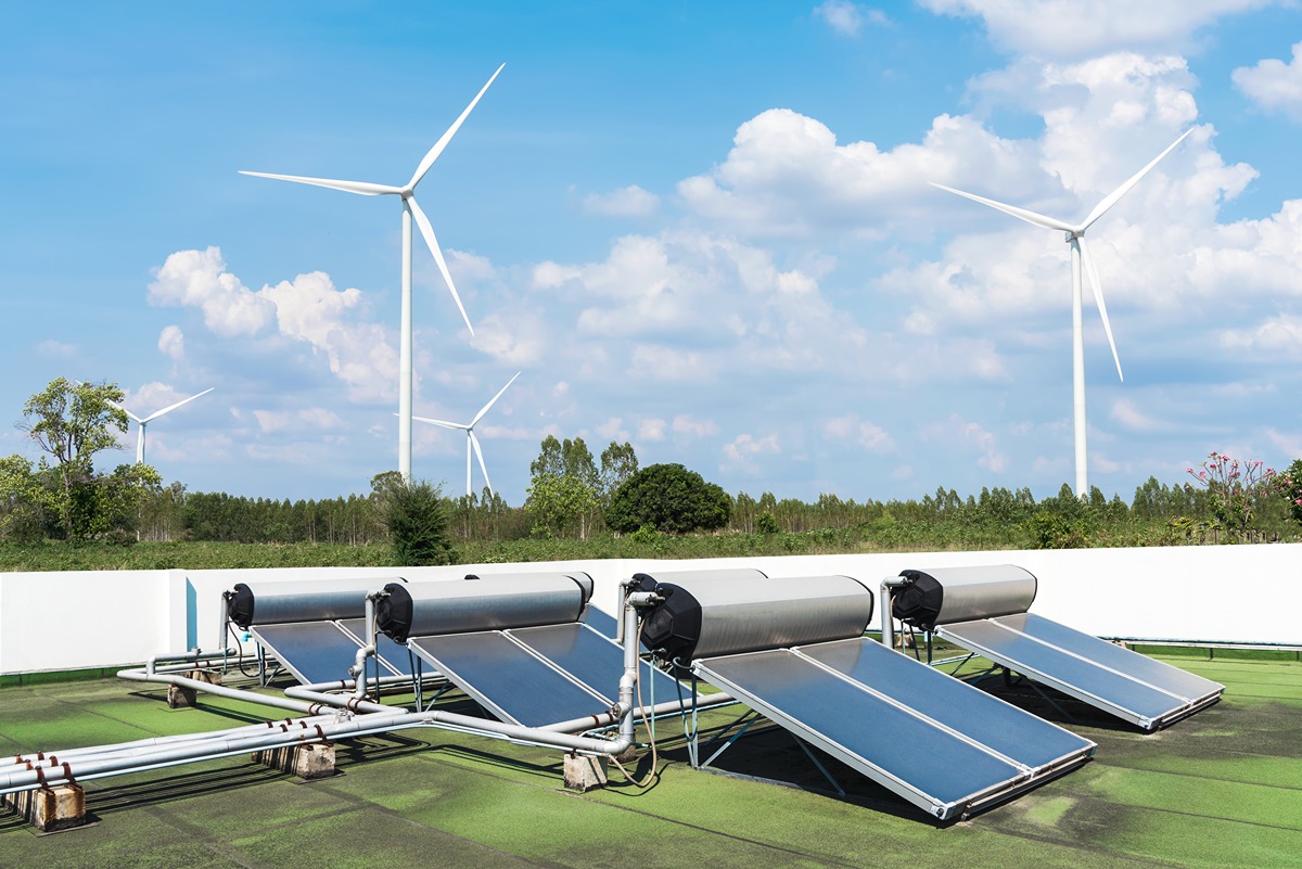 Image showcasing clean energy technologies with solar thermal panels installed on a green rooftop in the foreground and multiple wind turbines spinning in the background against a blue sky with scattered clouds. Trees line the horizon, emphasizing the integration of renewable energy sources in a natural setting.
