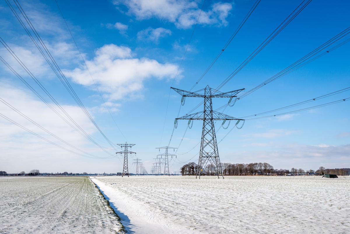 A winter landscape featuring multiple high-voltage power transmission towers and lines stretching across a snow-covered field under a partly cloudy blue sky.