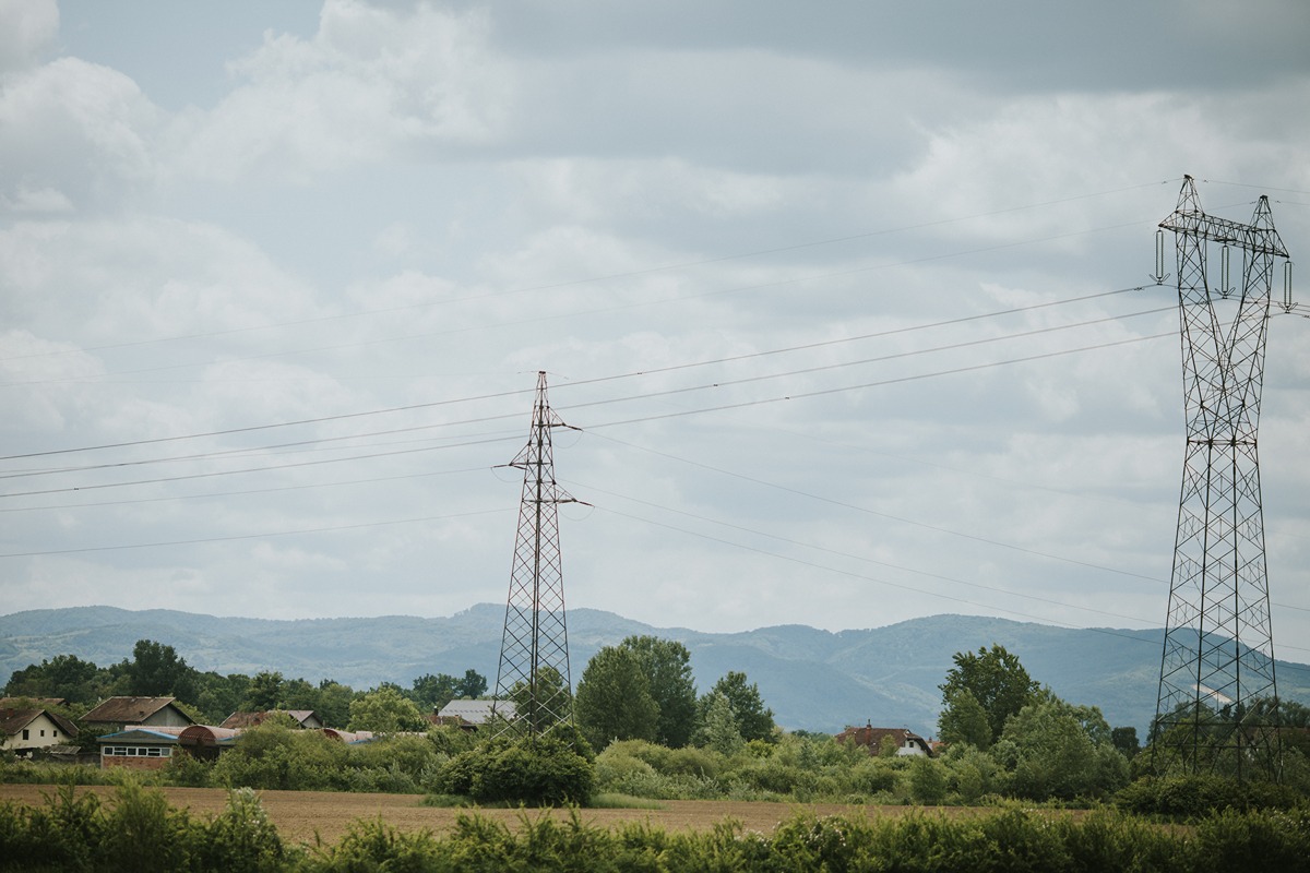 Two high-voltage transmission towers connected by power lines stand over a rural landscape with scattered houses, trees, and distant hills under a cloudy sky. The image represents infrastructure relevant to a large-scale 765-kilovolt transmission project.