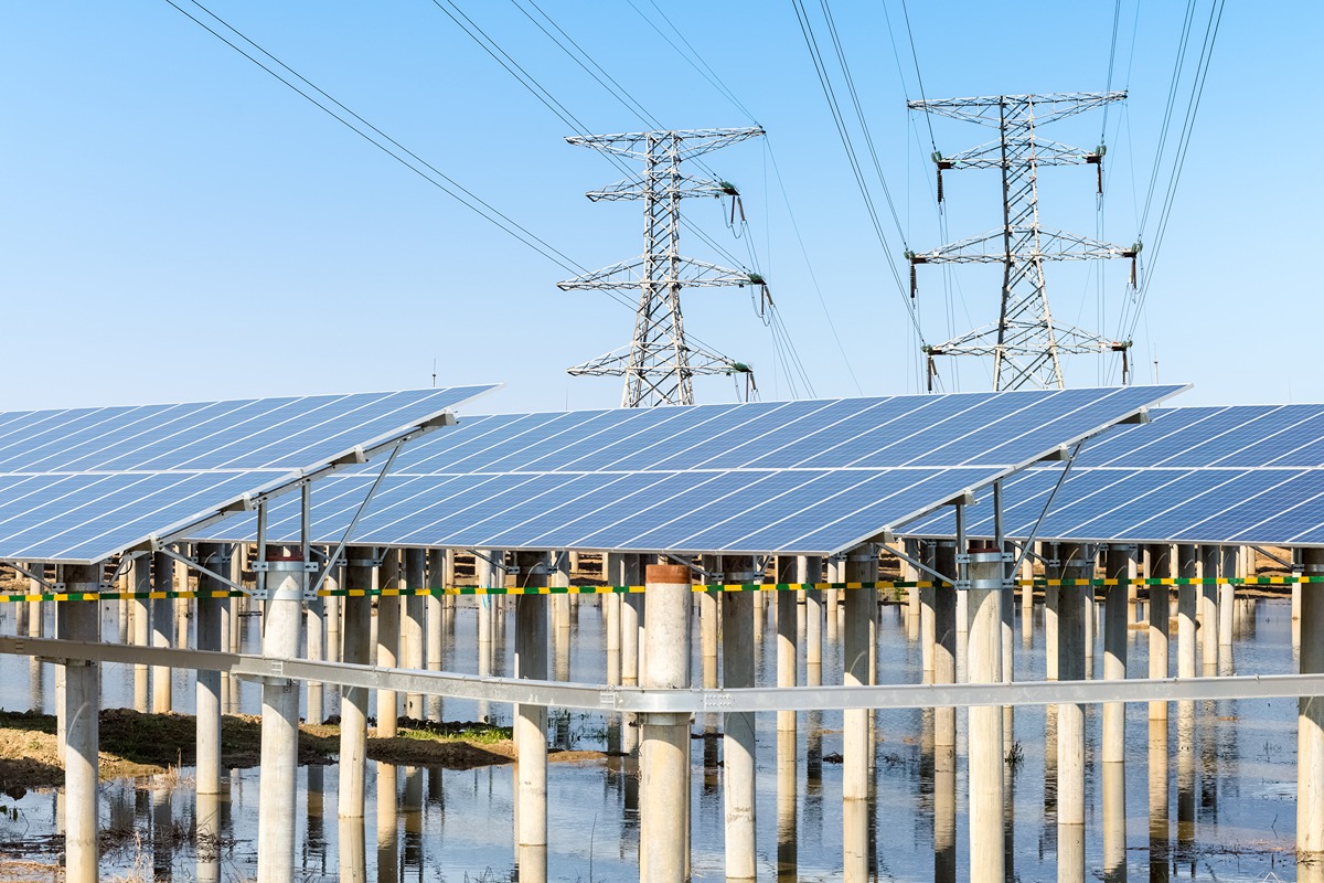 Elevated solar panels installed over a flooded or wetland area, with large electrical transmission towers and power lines in the background, illustrating renewable energy infrastructure expansion.
